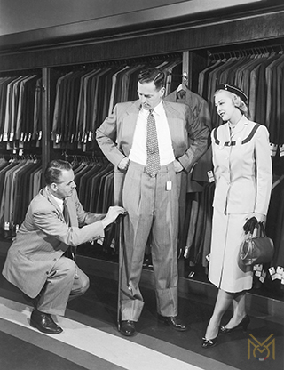 A tailor kneeling to take a client's trouser measurements.