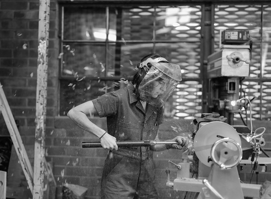 A craftsperson shaping a piece on a lathe behind a face shield - hands-on precision learned through practice.