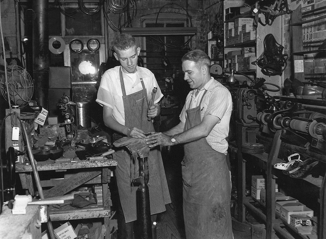 A shoemaker showing work to a younger colleague in a crowded workshop - craft passed on through close attention and real practice.
